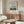 May include: Henri Rousseau vintage art poster displayed above wooden sideboard in minimalist living room. Cream-colored print features pastoral scene with cow, haystacks, and countryside landscape in muted greens and blues. Modern white sculptural chair and glass coffee table complete the Scandinavian-style interior with natural wood tones and soft lighting.