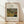 May include: Framed poster print leaning against white wall on wooden floor. Features Henri Rousseau's jungle painting with lush green tropical foliage, orange tiger partially visible among dense vegetation. Text reads "TIGER IN A TROPICAL" above artwork, "1891 FAMOUS PAINTING Henri Rousseau" below. Natural wood frame with cream matting. Soft window light creates diagonal shadows across white textured wall.