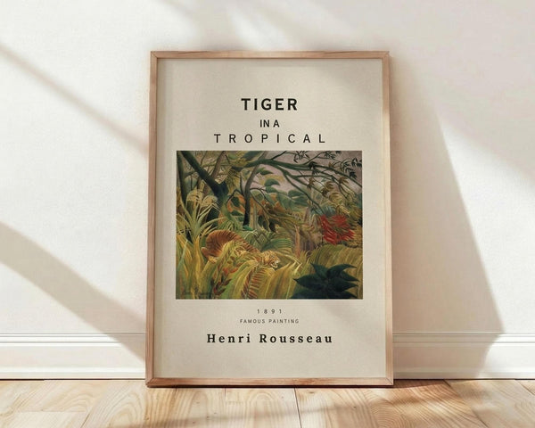 May include: Framed poster print leaning against white wall on wooden floor. Features Henri Rousseau's jungle painting with lush green tropical foliage, orange tiger partially visible among dense vegetation. Text reads "TIGER IN A TROPICAL" above artwork, "1891 FAMOUS PAINTING Henri Rousseau" below. Natural wood frame with cream matting. Soft window light creates diagonal shadows across white textured wall.