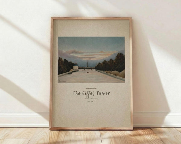 May include: Framed art print in light wood frame leaning against white wall on wooden floor. Print shows Henri Rousseau painting of wide tree-lined avenue leading toward distant Eiffel Tower under pale evening sky. Soft natural lighting creates diagonal shadows across the scene.