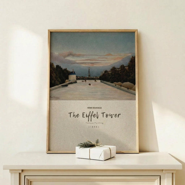 May include: Framed art print leaning against white wall on light wood surface, showing river scene with Eiffel Tower in distance, trees lining waterway, muted blue and pink sky. Small white wrapped gift with twine and greenery sprig sits in front of natural wood frame.