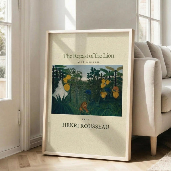 May include: Framed museum-style poster featuring Henri Rousseau's jungle painting with lush green foliage, yellow flowers, and a resting lion in dense vegetation. The print has cream-colored borders with black text identifying the artwork, artist, date 1907, and MET Museum. Light wood frame leans against white wall next to beige upholstered chair in bright, airy room with tall windows and natural light casting soft shadows on hardwood floors.