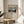 May include: Framed art print displayed on round wooden side table next to beige upholstered chair. Print shows riverside scene with Eiffel Tower in distance, rendered in muted blues and earth tones with cream matting. Natural lighting creates soft shadows across neutral-toned room setting with white walls and plant leaves visible at edge of frame.