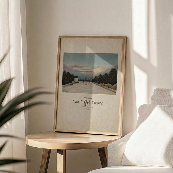 May include: Framed art print displayed on round wooden side table next to beige upholstered chair. Print shows riverside scene with Eiffel Tower in distance, rendered in muted blues and earth tones with cream matting. Natural lighting creates soft shadows across neutral-toned room setting with white walls and plant leaves visible at edge of frame.