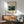 May include: Framed museum poster featuring tropical jungle scene with lush green foliage, yellow fruit, small lion figure, displayed above wooden sideboard with table lamp in modern living room with sage green armchair and natural lighting casting shadows on white wall.