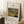 May include: Framed poster featuring Henri Rousseau's jungle painting with tiger hidden among lush green tropical foliage, displayed on white textured rug against white wall corner. Light wooden frame contains vintage-style museum poster layout with cream background and black text. Scene includes wooden side table with white coffee cup and saucer, cozy beige throw blanket visible at edge, soft natural lighting creating gentle shadows.