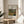 May include: Framed art print displaying Henri Rousseau's tropical jungle painting with dense green foliage and hidden tiger, featuring white mat with black text reading "TIGER IN A TROPICAL" and "Henri Rousseau" below. Thin gold frame rests on light wood round side table next to beige upholstered chair with plant leaves casting shadows on white wall in bright, minimalist room setting.