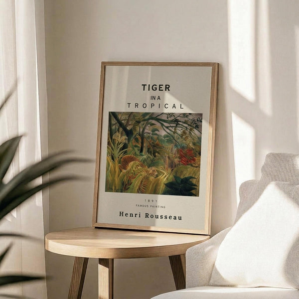 May include: Framed art print displaying Henri Rousseau's tropical jungle painting with dense green foliage and hidden tiger, featuring white mat with black text reading "TIGER IN A TROPICAL" and "Henri Rousseau" below. Thin gold frame rests on light wood round side table next to beige upholstered chair with plant leaves casting shadows on white wall in bright, minimalist room setting.