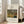 May include: Framed art print displayed in bright living room setting, leaning against white wall next to beige upholstered chair. Print features lush tropical jungle scene with dense green foliage, palm fronds, and orange tiger partially hidden among vegetation. White matted poster with black text above and below central artwork image, housed in natural wood frame. Sunlight streams through tall windows onto hardwood floors.