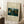 May include: Framed museum poster print of Henri Rousseau's jungle painting featuring a lion resting among lush tropical foliage with yellow flowers and dense green vegetation. The cream-colored poster displays "The Repast of the Lion" with "MET Museum" and "1907 HENRI ROUSSEAU" text. Light wooden frame leans against white wall corner near cozy seating area with coffee cup on wooden block side table and soft textured throw blanket.