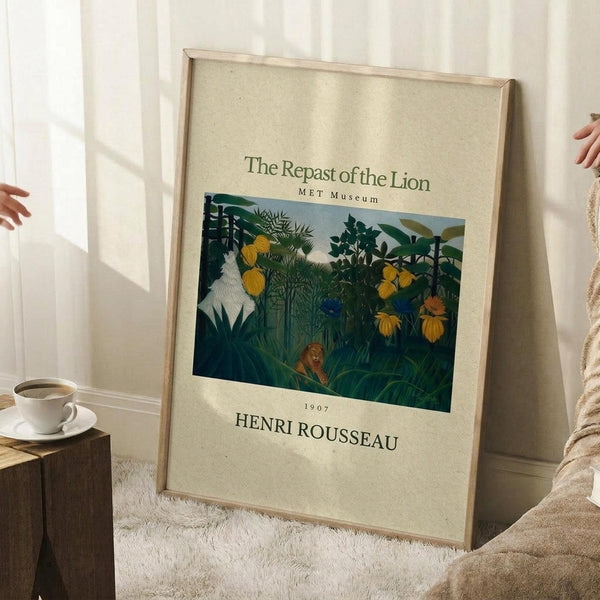 May include: Framed museum poster print of Henri Rousseau's jungle painting featuring a lion resting among lush tropical foliage with yellow flowers and dense green vegetation. The cream-colored poster displays "The Repast of the Lion" with "MET Museum" and "1907 HENRI ROUSSEAU" text. Light wooden frame leans against white wall corner near cozy seating area with coffee cup on wooden block side table and soft textured throw blanket.