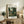 May include: Framed botanical art print featuring lush jungle foliage in deep greens with tropical plants and flowers, displayed on weathered wood sideboard alongside fern plant, ceramic vase, stacked books, and throw pillows in neutral living room setting with cream walls