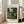 May include: Framed art print of a lush tropical jungle scene with dense green foliage, palm fronds, and exotic plants in muted greens and browns. The poster features gallery-style text at top and bottom on cream background. Displayed in light wooden frame leaning against white wall next to beige upholstered chair in bright, airy room with tall windows and hardwood floors.