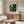 May include: Framed poster of Henri Rousseau's jungle painting featuring lush green tropical foliage and plants displayed above a natural wood sideboard with sliding doors. Modern living room setting with cream upholstered chair, glass-top oval coffee table, and minimalist white table lamp. Neutral beige walls with natural lighting from window.