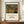 May include: Framed art print featuring Henri Rousseau's jungle painting with a tiger hidden among lush green tropical foliage and palm fronds during a storm. The poster has cream-colored text below the artwork identifying the title and artist. Displayed on a wooden sideboard beside a white ceramic vase and stacked books in an elegant room with cream walls and white molding.