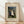 May include: Framed art print of Henri Rousseau's "The Wedding Party" painting from 1905, showing a bride in white dress surrounded by wedding guests in dark formal attire standing among tropical foliage with a small black dog at their feet. The print features cream matting in a light wooden frame, displayed on wooden flooring against a white wall with soft natural lighting creating diagonal shadows.