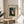 May include: Framed poster print of Henri Rousseau's The Wedding Party painting displayed on a round wooden side table next to a beige upholstered chair. The artwork shows a group wedding portrait with people in formal dark clothing and a bride in white, set against tropical foliage. Natural sunlight creates soft shadows across the neutral-toned room with white walls.