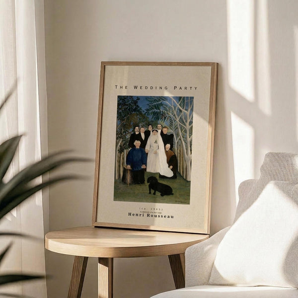 May include: Framed poster print of Henri Rousseau's The Wedding Party painting displayed on a round wooden side table next to a beige upholstered chair. The artwork shows a group wedding portrait with people in formal dark clothing and a bride in white, set against tropical foliage. Natural sunlight creates soft shadows across the neutral-toned room with white walls.