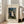May include: Framed art print leaning against beige sofa in bright living room with white walls and large windows. Print shows naive-style painting of wedding group with bride in white dress, men in dark suits, and black dog in forest setting with leafy trees. Cream matting surrounds colorful artwork in light wood frame on wooden floor.