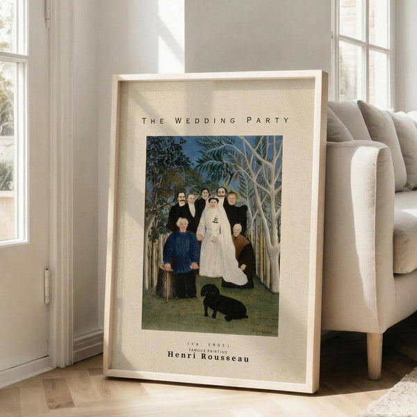 May include: Framed art print leaning against beige sofa in bright living room with white walls and large windows. Print shows naive-style painting of wedding group with bride in white dress, men in dark suits, and black dog in forest setting with leafy trees. Cream matting surrounds colorful artwork in light wood frame on wooden floor.