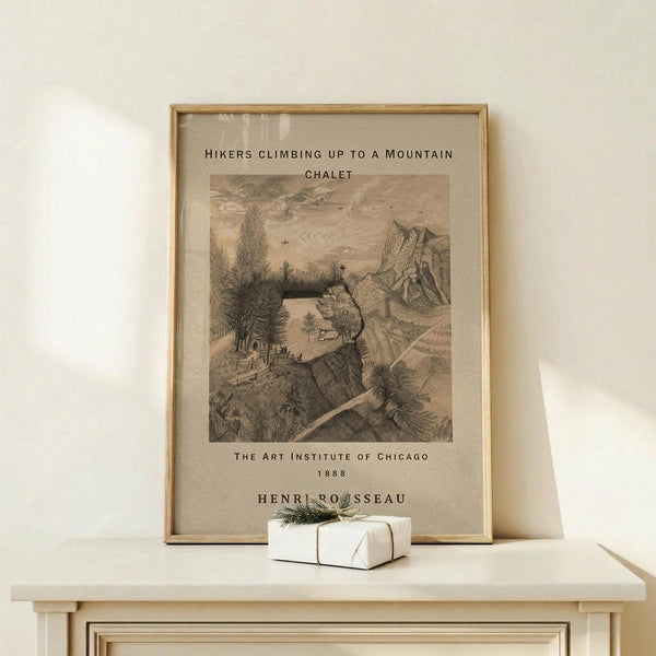 May include: Framed vintage art print showing hikers climbing toward a mountain chalet, rendered in sepia-toned etching style with detailed landscape scenery. Light wood frame displays the artwork on a pale wooden surface beside a small white gift box with greenery. Cream colored wall background creates minimal, natural styling.