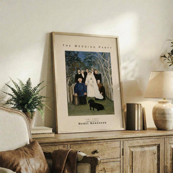 May include: Framed art print featuring Henri Rousseau's "The Wedding Party" painting displayed on a rustic wooden sideboard. The vintage-style poster shows a wedding scene with figures in dark clothing and a bride in white, set in a lush jungle landscape with tropical plants and a small black dog. The print has cream matting and light wood frame, styled with fern plant, ceramic vase, books, and neutral home decor in warm natural lighting.