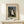 May include: Framed art print featuring Henri Rousseau's primitive style painting of a wedding party in a forest setting. The artwork shows figures in formal black attire surrounding a bride in white, with a black dog at their feet and leafy trees in the background. The print has cream matting with black text above and below the image. A small wrapped gift with twine and greenery sits on a light wood console table beneath the frame against a white wall.