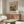 May include: Henri Rousseau vintage art print featuring hikers climbing toward a mountain chalet, displayed in thin wooden frame above walnut sideboard in modern neutral living room with white sculptural chair, glass oval coffee table, and warm natural lighting