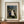 May include: Framed art print of Henri Rousseau's "The Wedding Party" painting displayed on wooden credenza beside white ceramic vase and books. Print shows bride in white dress surrounded by wedding party in dark formal wear, set in lush jungle landscape with tropical plants and small black dog. Cream matting with brown frame against white paneled wall.