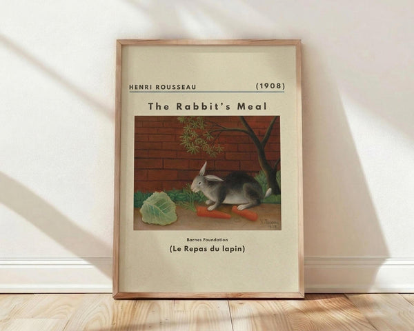 May include: Framed art print leaning against white wall on light wood floor. The poster shows a realistic painting of a gray and white rabbit sitting beside orange carrots and green cabbage leaves, with a brick wall and tree branch in the background. Cream-colored mat with black text identifying the artwork as Henri Rousseau's 1908 piece. Natural wood frame with visible grain, soft window light creating diagonal shadows across the white fabric backdrop.