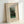 May include: Framed art print leaning against white wall showing Henri Rousseau's colorful painting of four figures in formal dress positioned among lush green trees and foliage. The cream matted poster features the artwork title and artist name in blue text. Natural wood frame with simple rectangular design creates clean, modern display against neutral background.