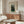 May include: Henri Rousseau art print in light wood frame hanging above walnut credenza in minimalist living room. Poster features two men in dark suits standing in lush green landscape with trees. Room has cream walls, white curved chair, glass oval coffee table, and modern brass lamp. Natural lighting creates warm, contemporary atmosphere showcasing the vintage botanical artwork.