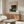 May include: A framed vintage-style art print featuring a jungle scene with a silhouetted figure among lush tropical plants and foliage under a moonlit sky, displayed above a mid-century modern walnut credenza in a minimalist living room with cream walls, white sculptural chair, glass oval coffee table, and modern table lamp.