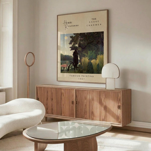 May include: A framed vintage-style art print featuring a jungle scene with a silhouetted figure among lush tropical plants and foliage under a moonlit sky, displayed above a mid-century modern walnut credenza in a minimalist living room with cream walls, white sculptural chair, glass oval coffee table, and modern table lamp.
