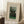 May include: Framed vintage art poster leaning against white paneled wall on white textured rug. Print shows two men in dark formal wear standing beneath green trees with two faces floating in blue sky above. Text reads "The Past and the Present, or Philosophical Thought" and "Henri Rousseau (1890)" in blue lettering on cream background. Natural wood frame, cozy room setting with coffee cup on wooden block nearby and beige blanket.