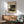 May include: Framed art print featuring Henri Rousseau's jungle scene with silhouetted figure among lush tropical foliage in green and yellow tones, hanging above wooden sideboard with white ceramic lamp, next to sage green upholstered armchair with walnut frame in modern living room with natural sunlight streaming across white walls.