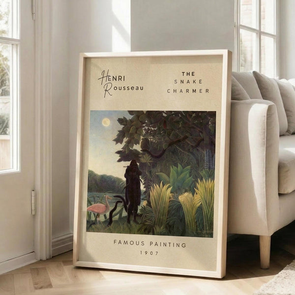 May include: Framed art print leaning against white wall beside beige sofa in bright living room with large windows. Print features tropical jungle scene with silhouetted figure among lush green foliage, flamingo, and moonlit sky. Cream colored mat with black text identifying the artwork and date 1907. Light wood frame, approximately 2-3 feet tall, positioned on hardwood floors with soft natural lighting.