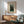 May include: Henri Rousseau poster print in wooden frame hanging on white wall above natural wood sideboard. Green upholstered mid-century chair with wooden arms sits nearby. The artwork shows two men in dark clothing standing outdoors with lush green foliage and trees. Warm sunlight streams across the minimalist room creating geometric shadows on the wall and furniture.