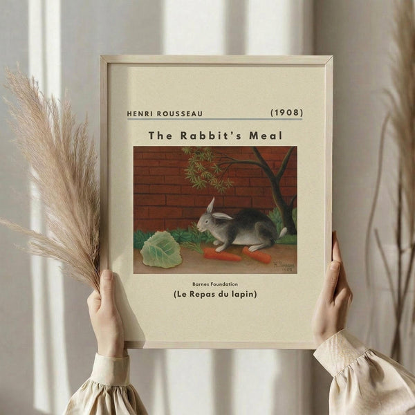 May include: Hands holding a framed art print featuring Henri Rousseau's painting of a gray and white rabbit sitting by orange carrots and green cabbage leaves against a brick wall background. The beige matted print is held alongside dried pampas grass stems in a softly lit room with cream curtains.