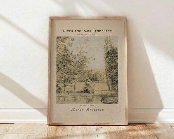 May include: Framed vintage landscape print in natural wood frame leaning against white wall on light wooden floor. Artwork shows sepia-toned drawing of rural scene with trees, small house, and water. Text reads "River and Park Landscape" at top and "Henri Rousseau" at bottom with Art Institute of Chicago attribution. Soft natural lighting creates shadows on textured white wall.