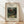 May include: Framed art print leaning against white wall on light wood floor. Print shows Henri Rousseau's jungle painting with dense green foliage, orange fruits, monkeys, and a red-orange parrot. Natural wood frame surrounds cream matting with black text above and below the artwork. Soft natural lighting creates diagonal shadows across the scene.