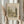 May include: Hands holding a framed vintage landscape print showing trees, water, and pastoral scenery in muted earth tones. Light wood frame against neutral background with dried pampas grass visible on left side. Person wearing cream-colored sweater with gathered cuffs.