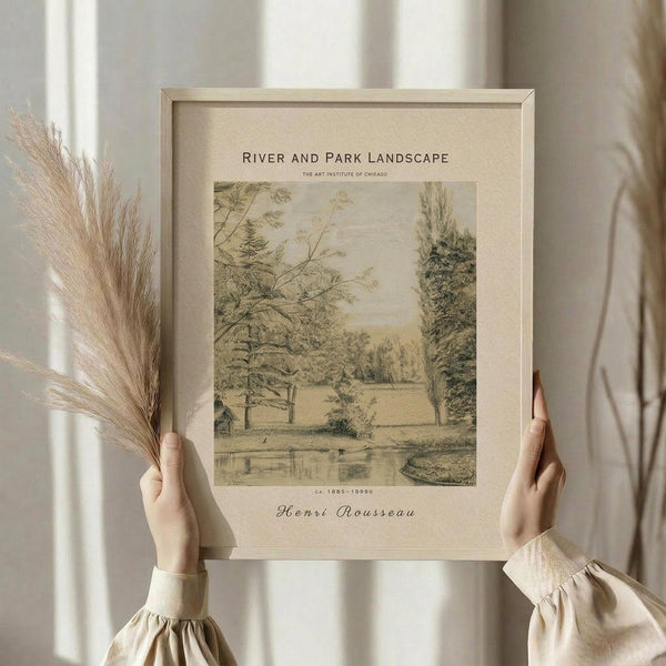 May include: Hands holding a framed vintage landscape print showing trees, water, and pastoral scenery in muted earth tones. Light wood frame against neutral background with dried pampas grass visible on left side. Person wearing cream-colored sweater with gathered cuffs.