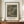 May include: Framed art print featuring Henri Rousseau's 1885 etching of riverside buildings and castle ruins in muted green and beige tones, displayed on wooden sideboard next to white ceramic vase and stacked books against cream wainscoted wall