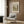 May include: Vintage landscape poster print with Henri Rousseau artwork displayed on wood paneled wall above beige upholstered armchair with geometric pillow. Print shows pastoral scene with trees, fields and bridge in muted green and cream tones within light wood frame. Modern reading nook with brass floor lamp and dark side table.