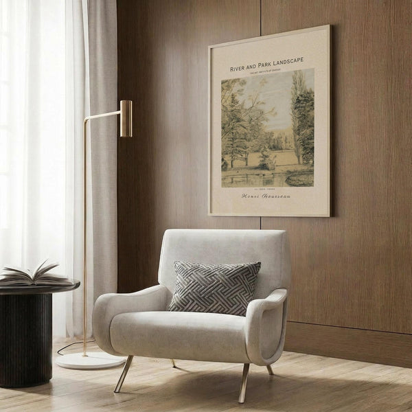 May include: Vintage landscape poster print with Henri Rousseau artwork displayed on wood paneled wall above beige upholstered armchair with geometric pillow. Print shows pastoral scene with trees, fields and bridge in muted green and cream tones within light wood frame. Modern reading nook with brass floor lamp and dark side table.