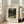 May include: Framed art print leaning against white wall next to beige sofa in bright living room. Print shows tropical jungle scene with monkeys and red parrot among lush green foliage, titled "Monkeys and Parrot in the Virgin Forest" by Henri Rousseau. Light wood frame with cream matting, natural lighting from windows creating shadows on hardwood floor.