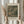 May include: Person in cream outfit holding framed art print showing muted green landscape scene with buildings and countryside, dried pampas grass in left hand, soft natural lighting by white curtained window, neutral beige wooden frame, vintage museum-style poster layout with artist signature and exhibition text below artwork