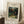 May include: Framed art print leaning against white wall showing tropical jungle scene with monkeys and red parrot among lush green foliage. Print has cream matting with black text identifying it as Henri Rousseau artwork from Barnes Foundation, dated ca. 1905-1906. Scene shows coffee cup on wooden surface and textured beige throw blanket nearby.