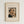 May include: Framed vintage-style art print displaying Henri Rousseau's self-portrait from 1903. Shows a balding man with dark hair, mustache, and formal black suit with bow tie beside an oil lamp. Cream colored background with brown text and wooden frame, mounted on white wall.